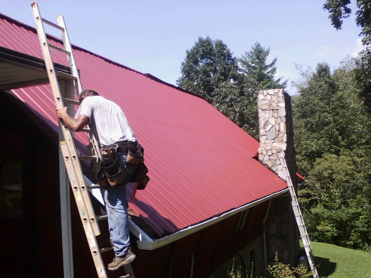 Roofer on a ladder beside a red metal roof and stone chimney — Soffit & Fascia Repair by Roofing Brothers in Beaver Meadows